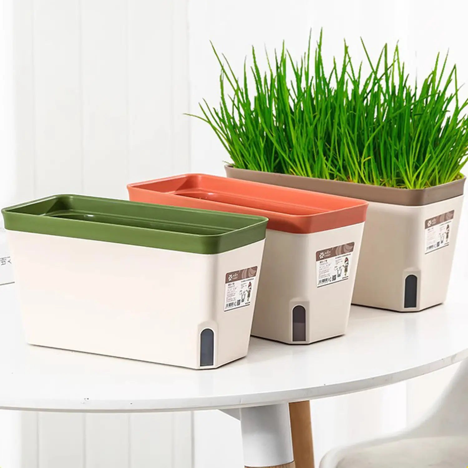 Set aus drei beigen rechteckigen Kunststoff-Blumentöpfen mit grünen, orangefarbenen und braunen Deckeln, mit automatischer Bewässerung und Baumwollstreifen für eine effiziente Pflanzenpflege.