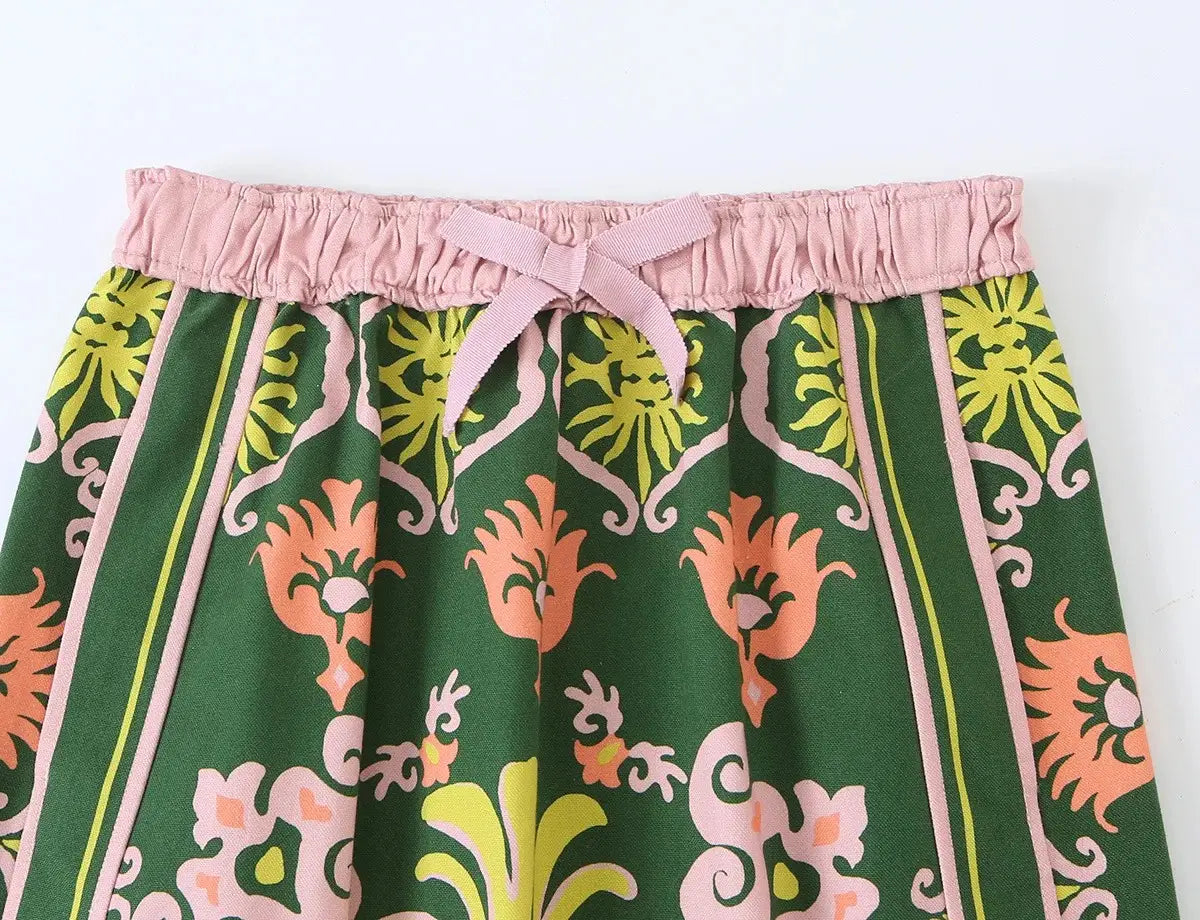 Ein grüner, hochgeschnittener Midi-Rock mit elastischem Bund in Pink und Schleife; mit kräftigen Korall-, Limettengrün- und blushrosa Blumenmustern auf dunkelgrünem Stoff aus dem floralen zweiteiligen Midi-Rock-Set im Boho-Outfit-Stil.