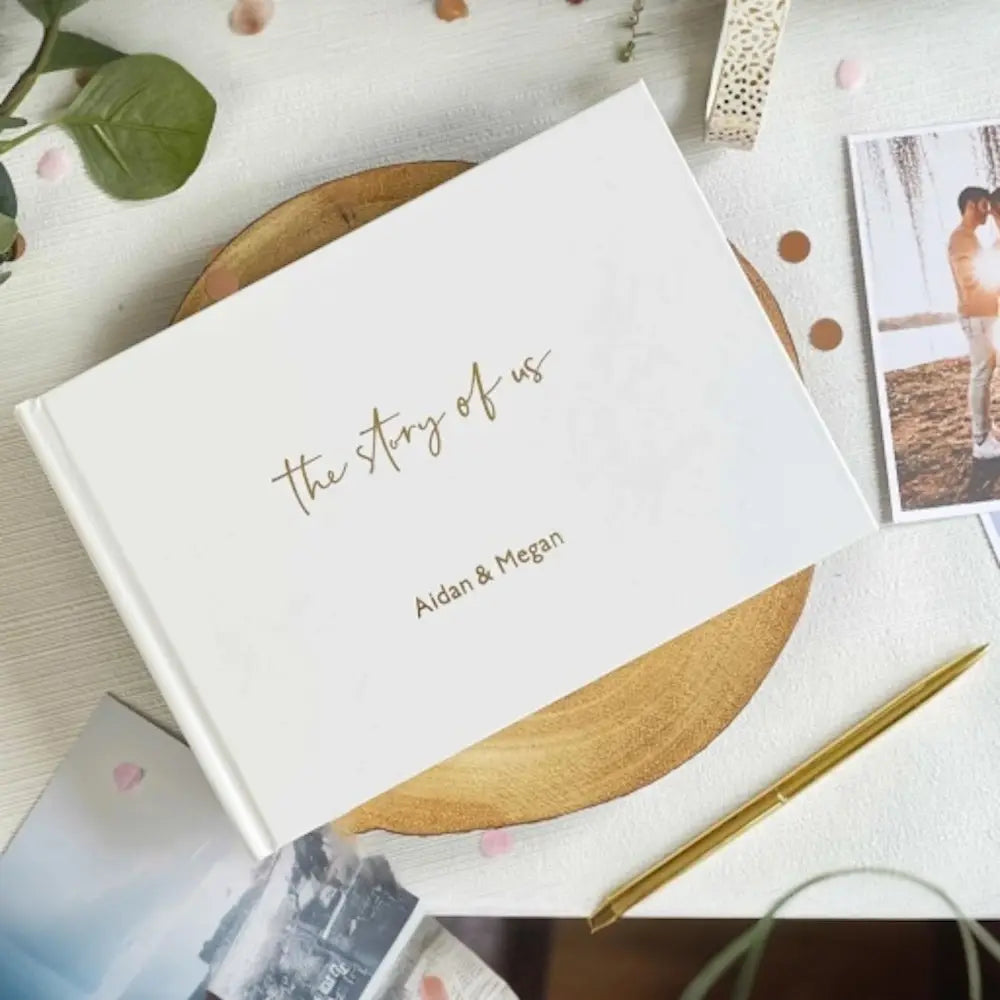 「the story of us」とエイダン、メーガンを金箔のレタリングであしらった、金箔文字の白いハードカバー写真アルバム。カスタム結婚式ゲストブックの記念品として個別にパーソナライズ。