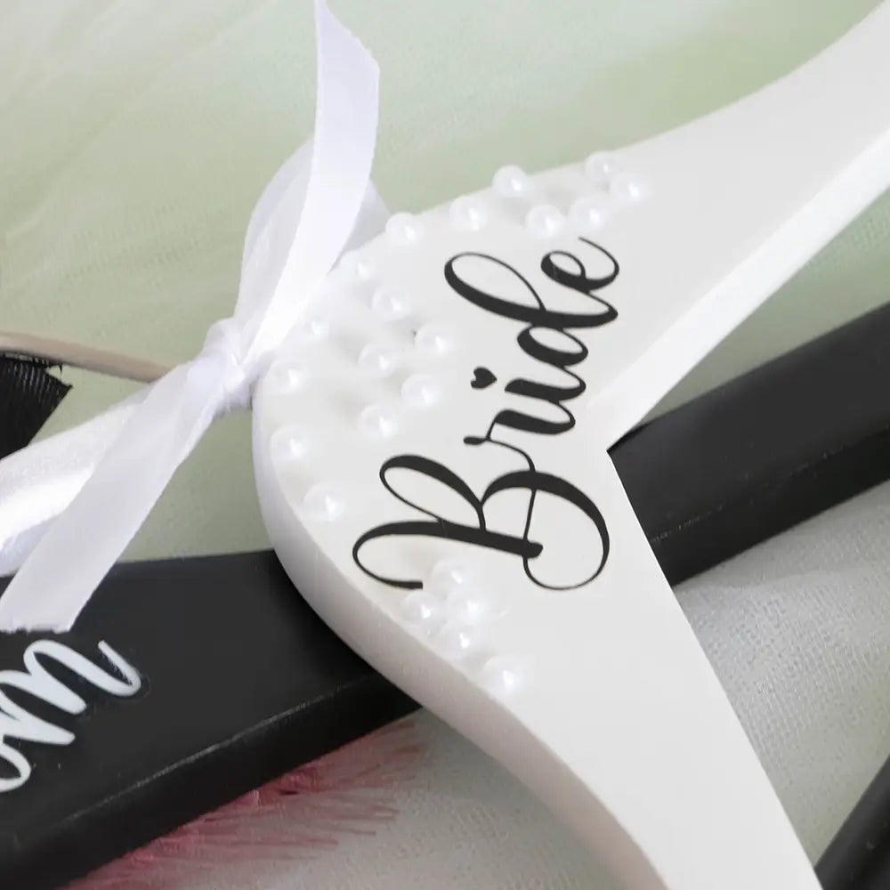 Cintre de mariage de la mariée en bois blanc, avec lettres cursives noires et de petites perles blanches le long du bord, provenant de l’ensemble de cintres de mariage perles pour la mariée et le marié.