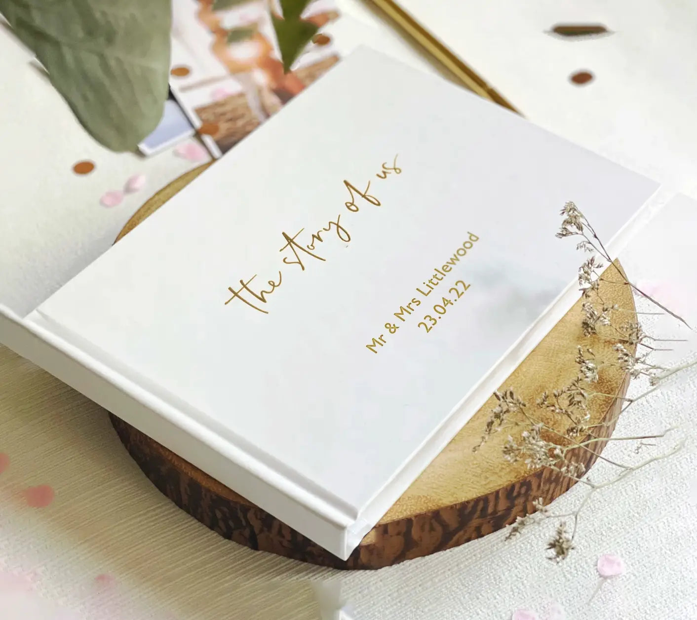 木のスライスの上に「the story of us」と「Mr and Mrs littlewood 23.04.22」をのせ、ゴールド箔レタリングでパーソナライズされた白いカスタム結婚式ゲストブック。