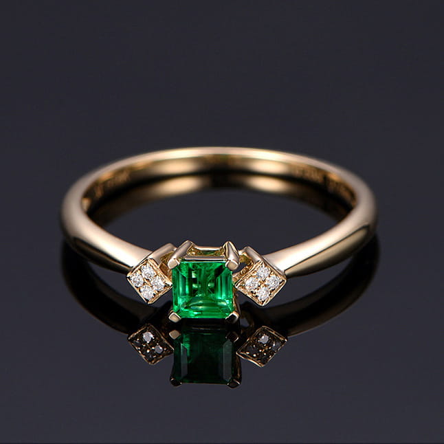 Ein Verlobungsring aus 18k Gold mit Smaragd und Diamanten: mit einem zentralen smaragdschliffartigen, grünen Edelstein, flankiert von zwei kleineren Diamant-Ansammlungen.