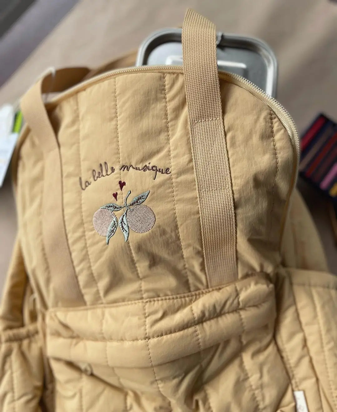 Beige gesteppten Wickeltaschen-/Tote Bag für Kinder aus wasserfestem Material mit besticktem Text „la belle musique“ und einem Pfirsich-Design, geeignet für die Schule.