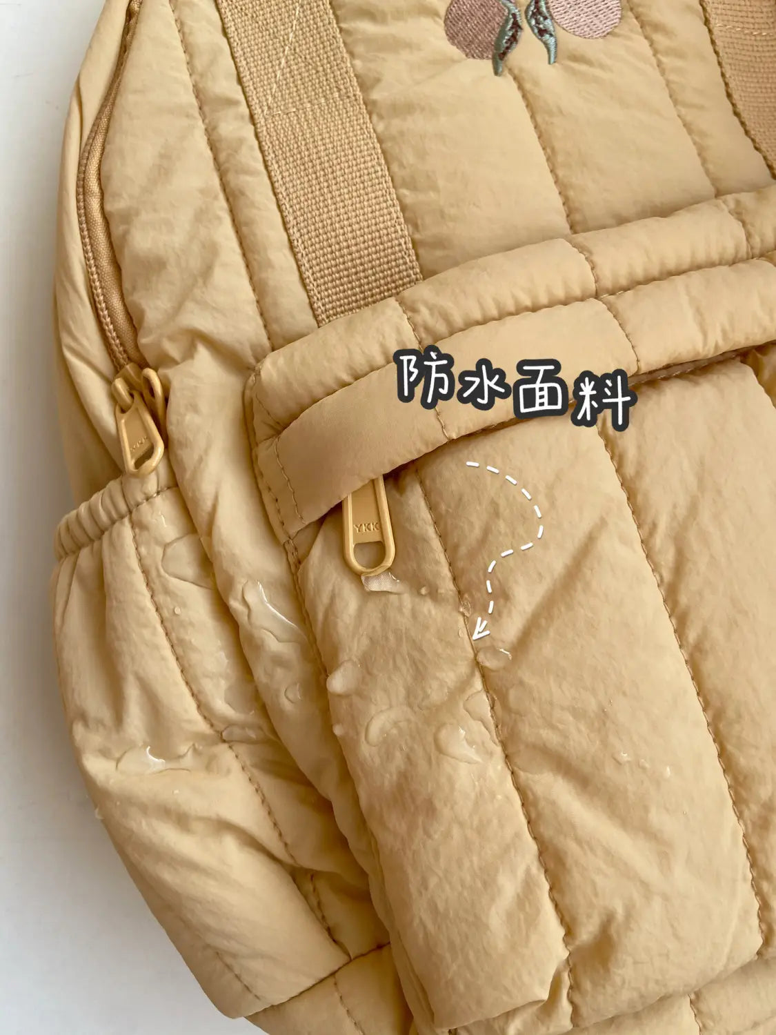 Ein beiger gesteppten Kinder-Rucksack aus wasserabweisendem Stoff mit bestickten Pfirsich-Applikationen und beigen Webband-Trägern, entworfen als wasserfester Schulrucksack.