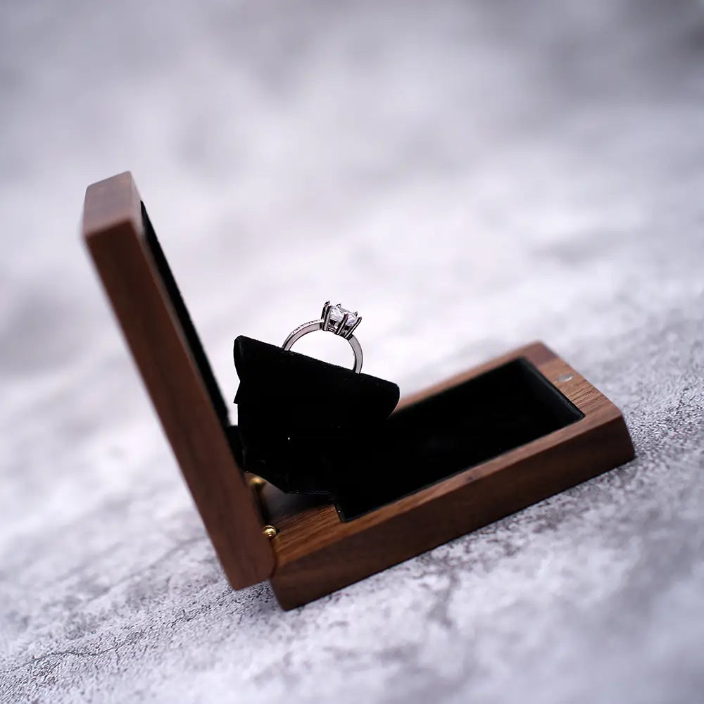 Ein silberner Verlobungsring mit einem zentralen Diamanten und vier Krappen, gebettet in einem dunklen Samtkissen, innerhalb einer geöffneten personalisierten Schmuckring-Schachtel aus Walnussholz zur Hochzeit.