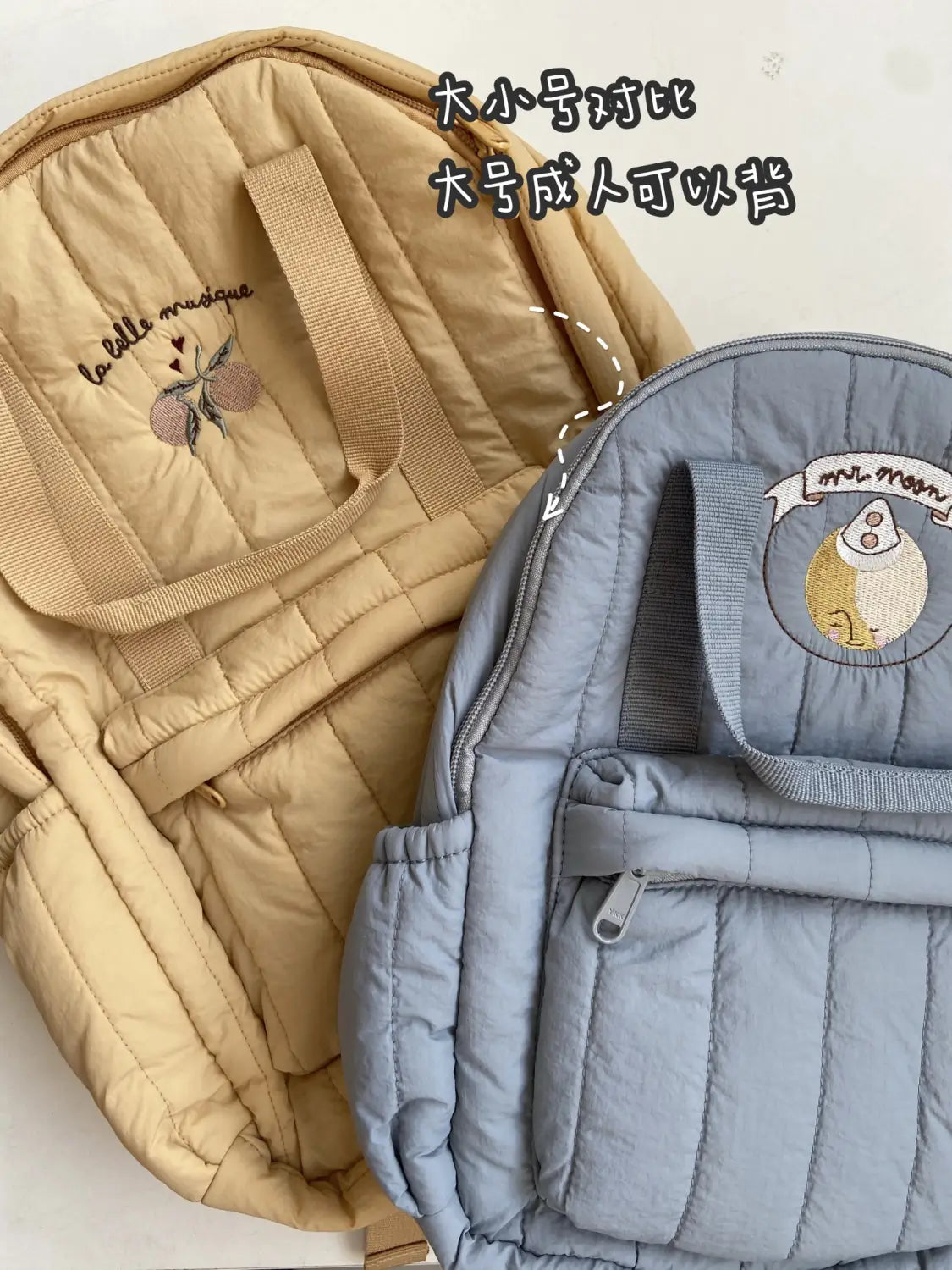 Ein gesteppten Rucksack für Kinder in Beige und Hellblau aus wasserfestem Material mit bestickten Motiven und kleinen Bannern mit den Aufschriften „la belle musique“ und „mammon“, geeignet für den Schulgebrauch