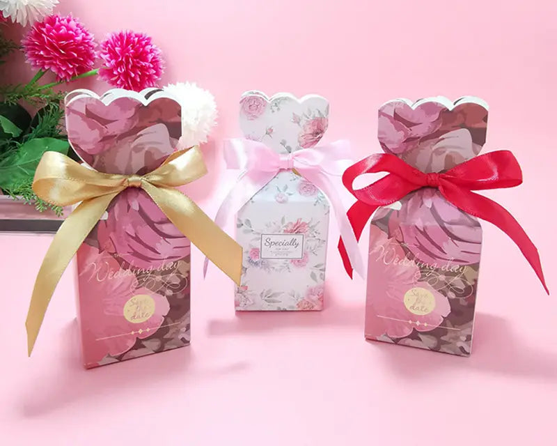 Drei Hochzeits-Gastgeschenkboxen mit Blumenmuster aus Satinband-Schleifen in Goldrosa und Rot, mit zarten Rosenmotiven auf den Papieroberflächen für eine elegante Geschenkpräsentation