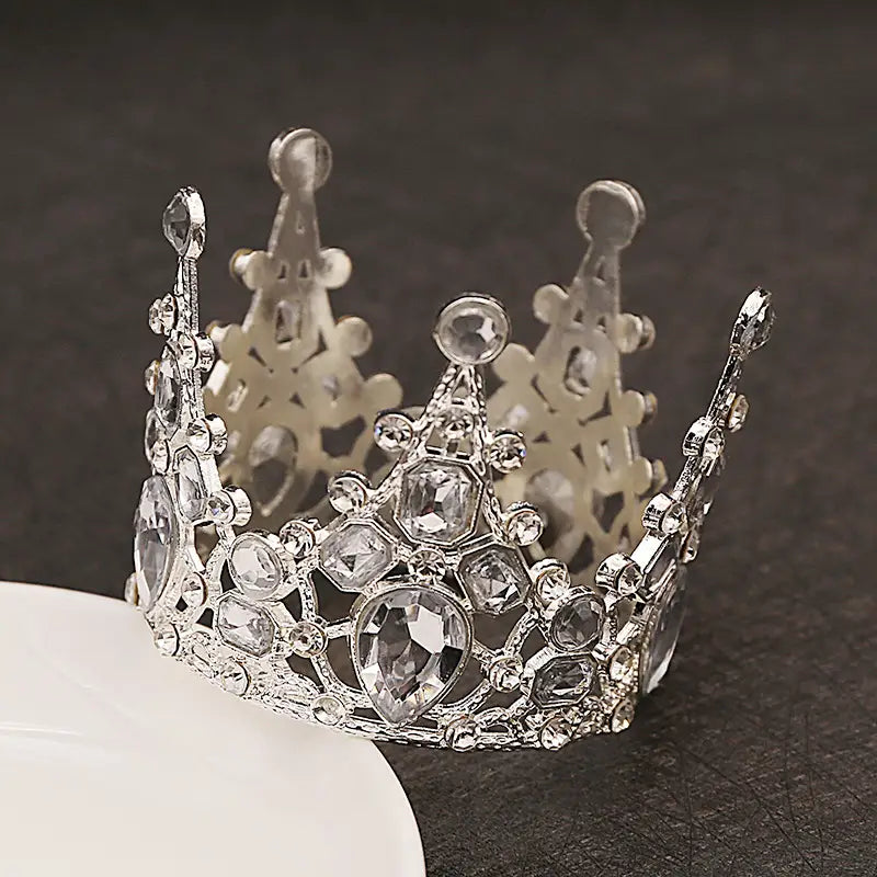Ein funkelnder silberner Mini-Kronen-Tortenaufsatz und eine Haar-Tiara, verziert mit klaren Kristallen und detaillierter, kunstvoller Filigranarbeit.