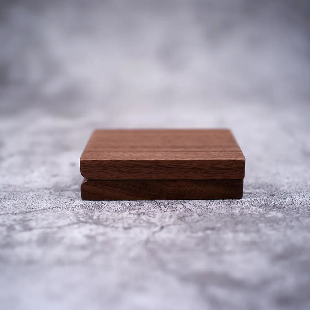 zwei eckige Untersetzer aus Holz in unterschiedlichen Walnusstönen, ordentlich aufgestapelt auf einer strukturierten Oberfläche, als Teil eines Sets für eine personalisierte Schmuckring-Schachtel aus Walnussholz zur Hochzeit.