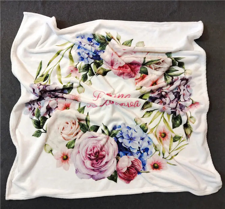 Eine weiche weiße, personalisierte Baby-Wickeldecke aus Fleece mit einem lebendigen Blumendesign aus pinken, blauen und violetten Rosen sowie Hortensien.