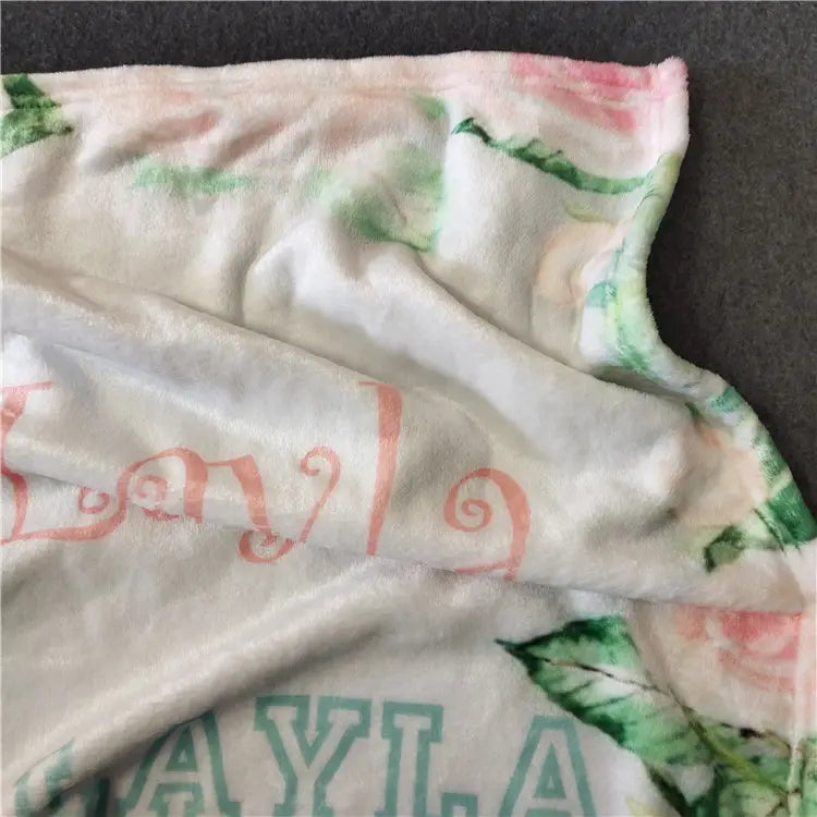 Eine personalisierte Baby-Wickeldecke aus Fleece mit weichem, plüschigem Material, einem rosa-grünen floralen Muster und dem Namen Layla, im Cursive-Stil aufgedruckt.