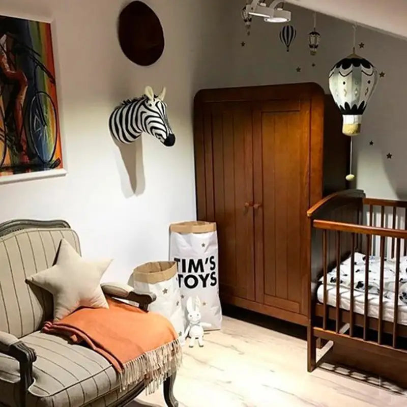 Ein hölzernes Babybett mit vertikalen Lamellen und einer geschwungenen Kopfplatte, mit weißer Bettwäsche mit schwarzen Mustern, neben einer 3D-Cartoon-Tierkopf-Wanddekoration angezeigt