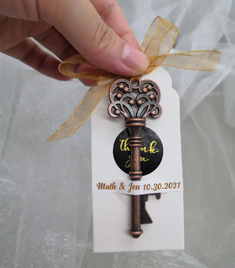 Encanto de llave antigua ornamentada color bronce, con detalles de filigrana intrincados, atado a una etiqueta blanca con cinta dorada como recuerdo abridor de botellas para bodas personalizado.