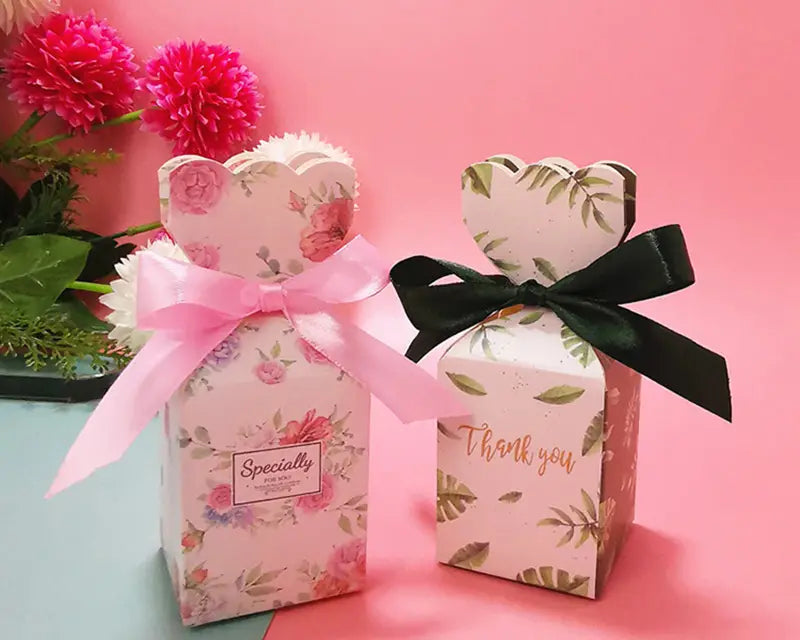 Zwei dekorative Hochzeits-Gastgeschenkboxen aus Papier mit herzförmigen Oberteilen, gebunden mit rosa- und schwarzfarbenen Bändern.