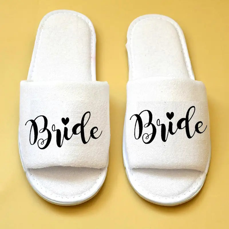 Weiße flauschige Schlupf-Slipper mit „Braut“ in schwarzer, geschwungener Handschrift und einem kleinen Herz über dem i für Brautparty-Slipper für Braut und Bräutigam als Hochzeits-Slipper.