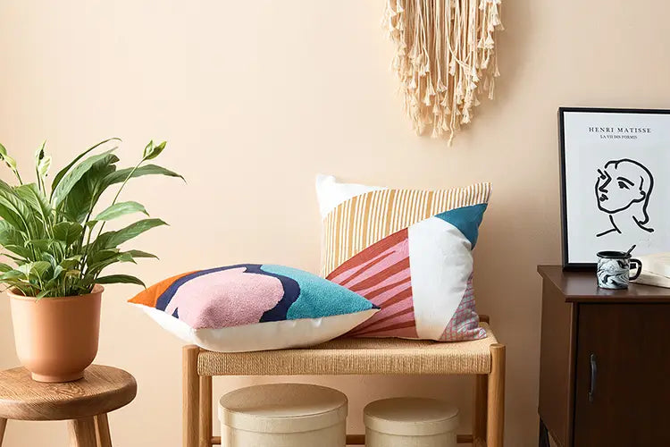 zwei bunte bestickte geometrische Kissenbezüge in den Größen 45x45 cm und 30x50 cm mit kräftigen Pink-, Blau-, Orange- und Weißmustern auf einer gewebten Holzbank