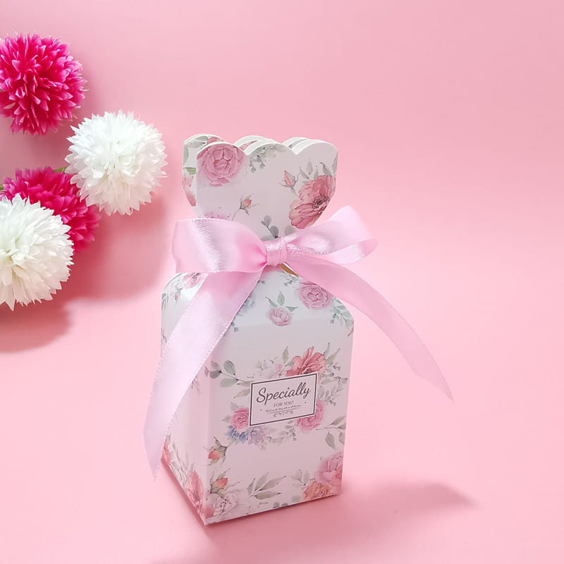 Eine zarte weiße Papier-Hochzeits-Gastgeschenkbox mit sanften rosa- und grünen Blumenelementen, gebunden mit einer Schleife aus Satinband.