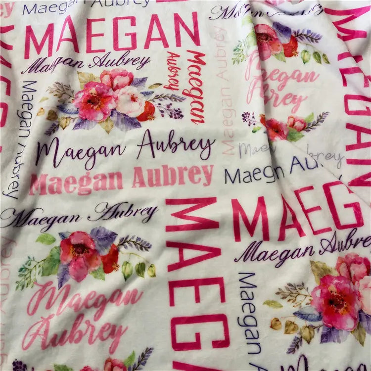 Personalisierte Baby-Wickeldecke aus weißem Fleece mit einem sich wiederholenden Muster des Namens maegan aubrey in verschiedenen Schriftarten und Farben, mit rosa- und lilafarbenen Blumen im Aquarellstil.