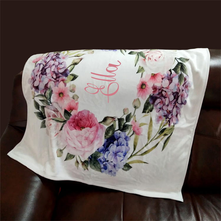 Eine weiße, personalisierte Baby-Wickeldecke aus Fleece mit einem lebendigen floralen Kranz aus pinken, violetten und blauen Blumen, der den Namen Ella von elegant in Schreibschrift umrahmt.