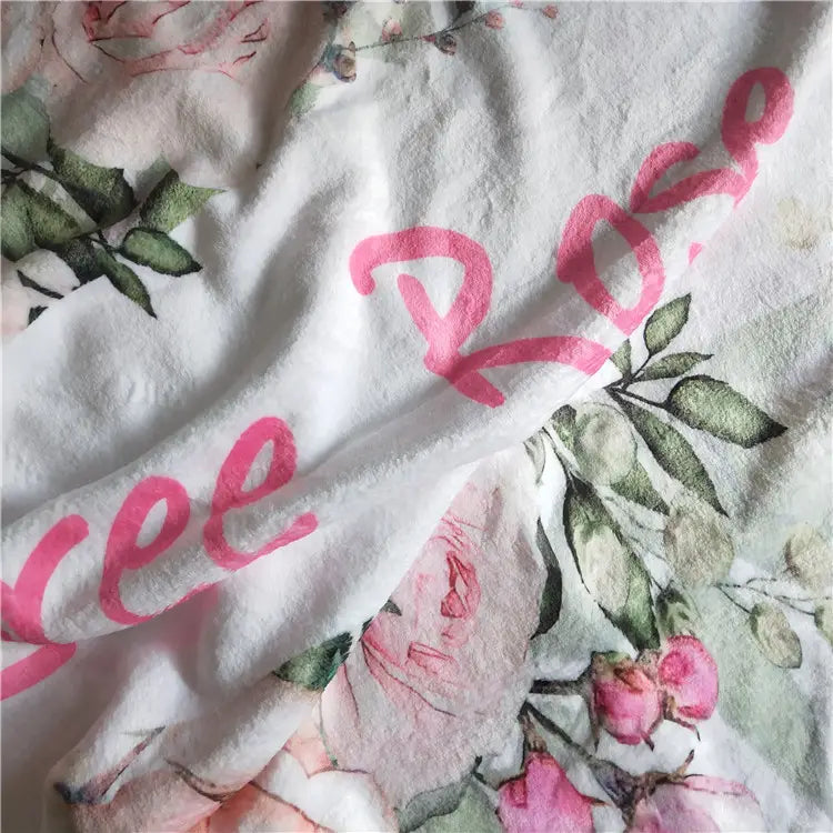 Personalisierte Baby-Wickeldecke aus Fleece in weißem Baumwollmaterial mit einem zarten floralen Muster aus pinken Rosen und grünen Blättern, akzentuiert durch kräftige pinkfarbene Schreibschrift.