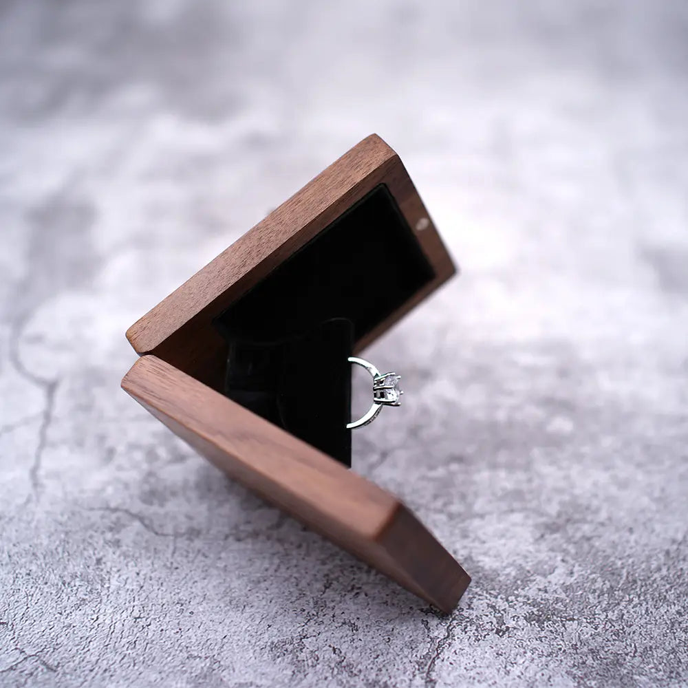 Ein silberner Ring mit einem klaren facettierten Edelstein, eingesetzt in einem minimalistischen Band, liegt in einer geöffneten personalisierten Schmuckring-Schachtel aus Walnussholz zur Hochzeit, ausgekleidet mit schwarzem Samt.