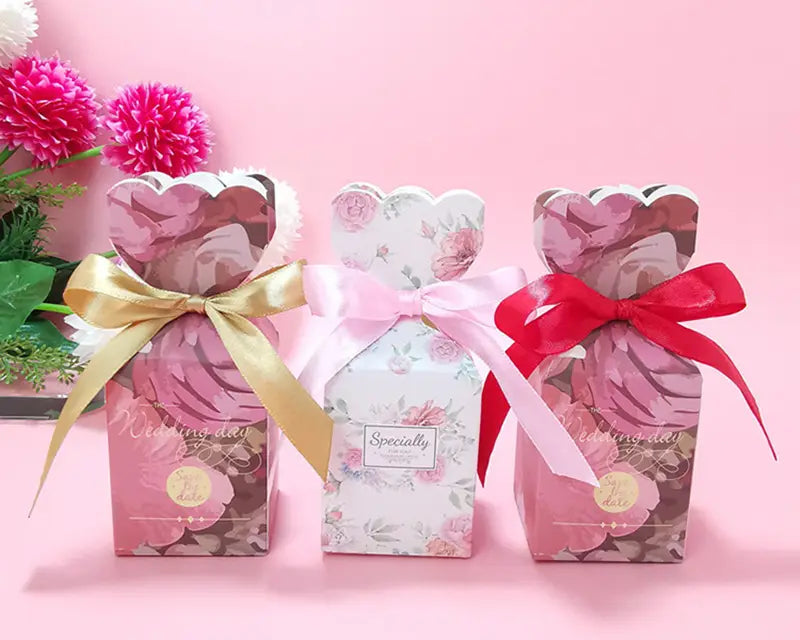Drei Blumige Hochzeits-Gastgeschenkboxen mit gewellten Oberkanten, gebunden mit Satinbändern in Goldrosa und Rot.