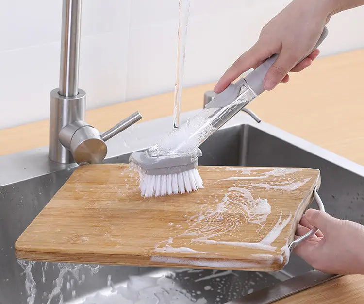 brosse en plastique grise et transparente pour distribution de savon, avec soies blanches et long manche, utilisée pour nettoyer une planche à découper en bois sous l’eau courante