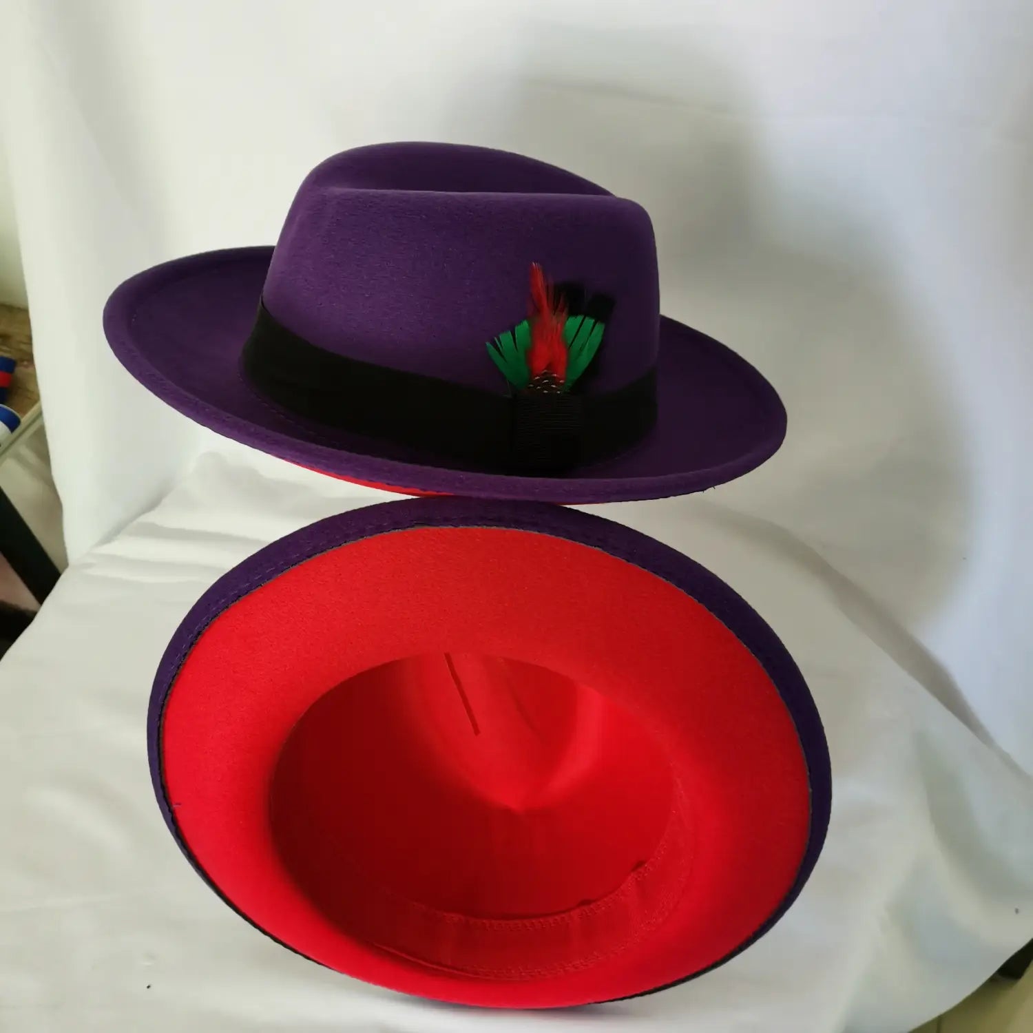 Unisex woolen felt fedora hat in purple with a black band and red and green feather accent featuring a vibrant red interior lining.