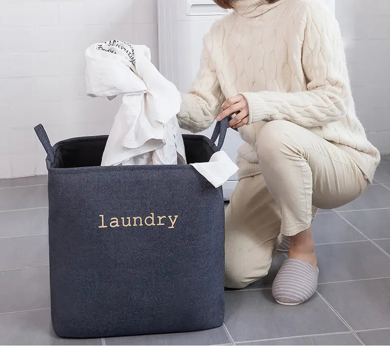 Dark blue fabric foldable laundry basket with laundry embroidered in gold thread designed as a storage basket for organizing clothes.