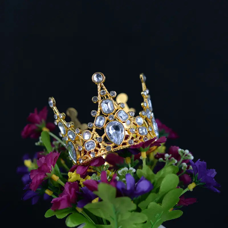 Ein funkelnder goldener Mini-Kronen-Tortenaufsatz und eine Haar-Tiara, verziert mit klaren facettierten Kristallen, die auf einem lebhaften Strauß aus lila, rosa und grünen Blumen platziert sind.