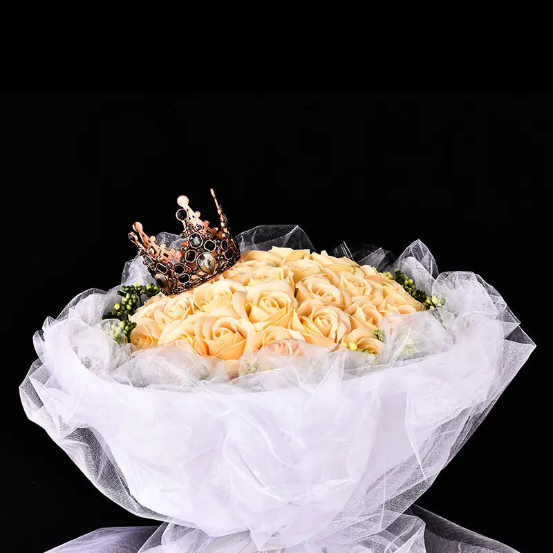 Ein Mini-Kronen-Tortenaufsatz und eine Haar-Tiara aus kunstvoll gearbeitetem, goldfarbenem Metall mit schwarzen Einlagen und einem zentralen Perlenbesatz, der auf einem Strauß blassgelber Rosen ruht, umwickelt mit transparentem weißem Tüll.