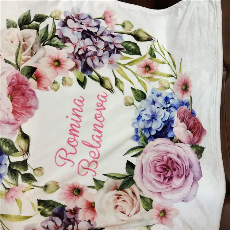 Eine weiße, personalisierte Baby-Wickeldecke aus Fleece mit einem lebendigen floralen Kranz aus pinken, violetten und blauen Blüten, umrahmt den Namen Romina Belanova in eleganter Schreibschrift.