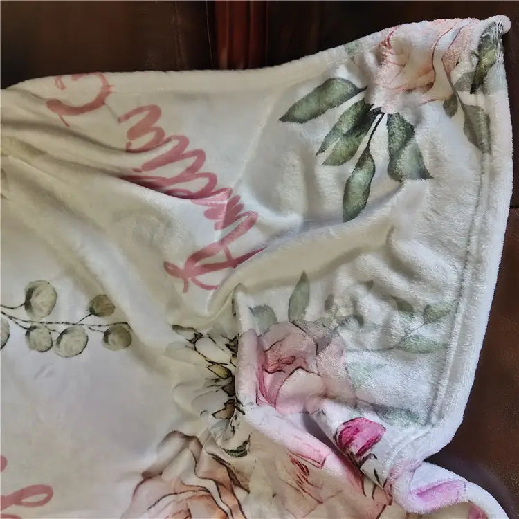 Eine weiche cremefarbene, personalisierte Baby-Wickeldecke aus Fleece mit einem zarten floralen Muster aus pinken Rosen, grünen Blättern und dezentem Cursive-Schriftzug in abgestimmten Rosatönen.