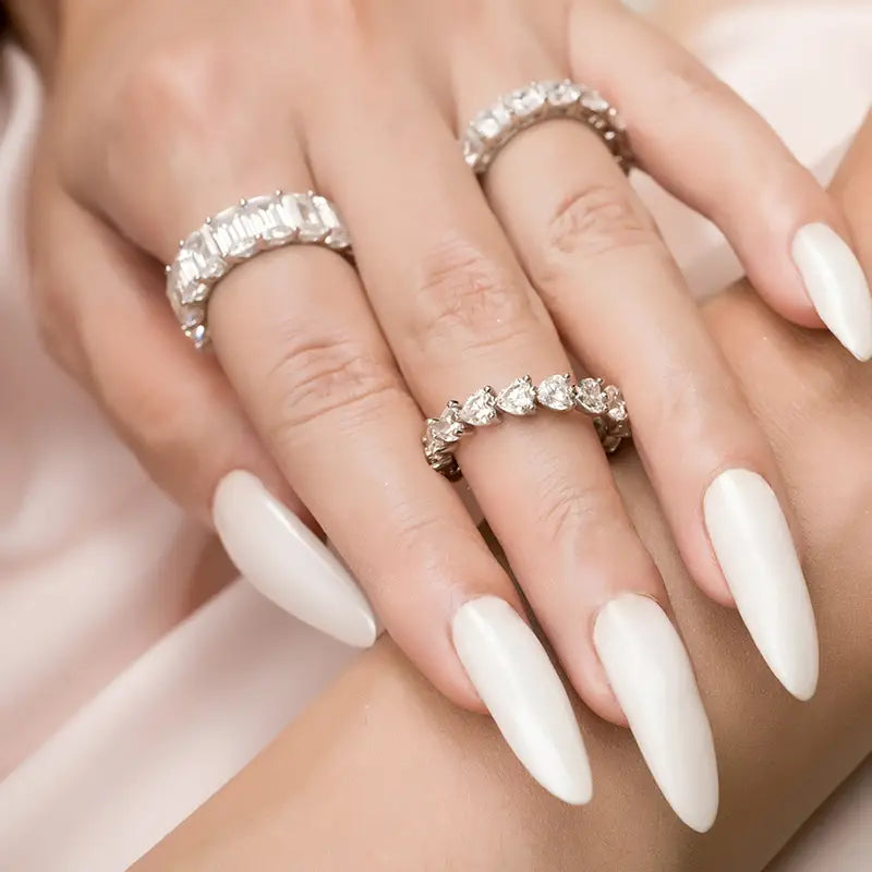 Une paire de mains aux ongles acryliques blancs longs, portant deux bagues d’éternité cœur en argent sterling distinctes, ornées de diamants.