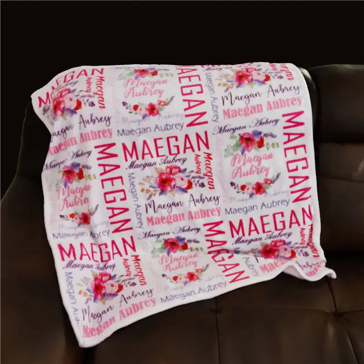 Eine weiche weiße, personalisierte Baby-Wickeldecke aus Fleece mit einem sich wiederholenden Muster des Namens Maegan Aubrey in kräftiger pinkfarbener und zarter Schrift sowie lebhaften floralen Illustrationen.