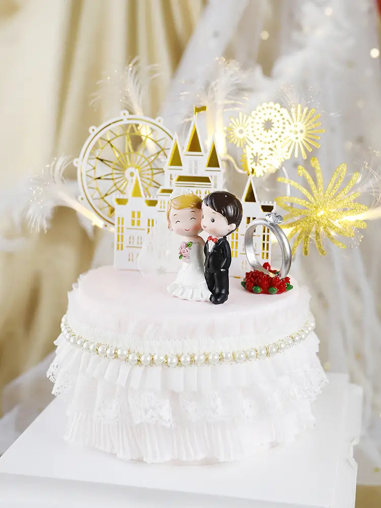 White wedding cake topper featuring bride and groom acrylic figurines with a gold castle and ferris wheel backdrop glittery golden fireworks accents and a silver ring on red berries.
