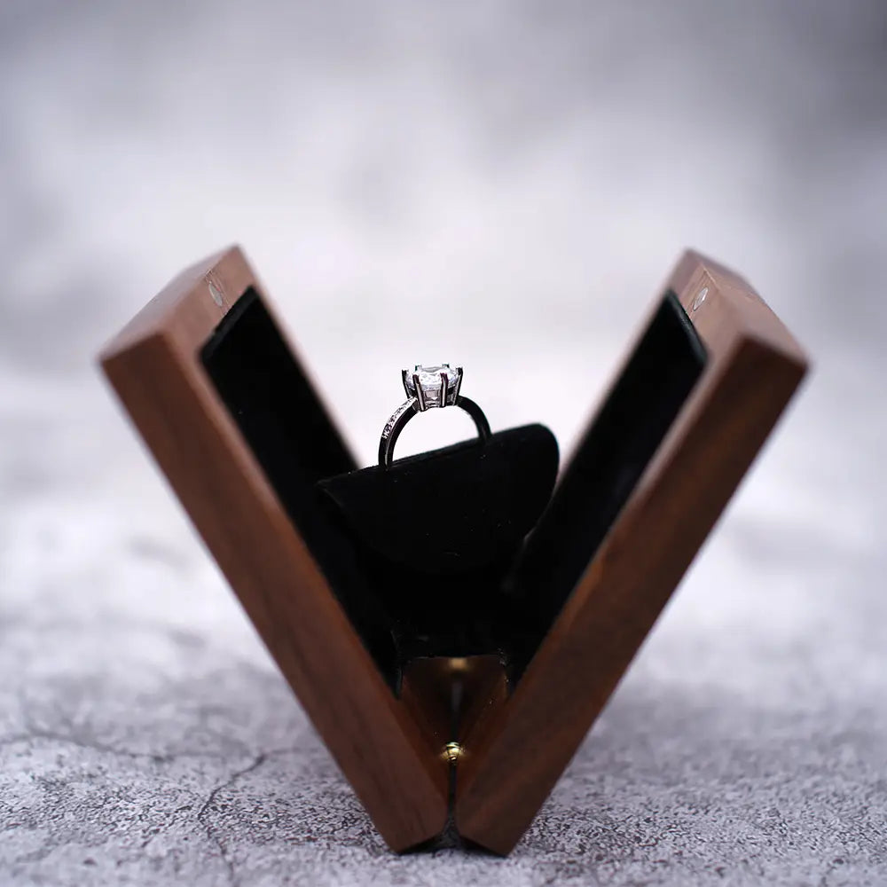 Ein eleganter silberfarbener Verlobungsring mit einem hervorstechenden zentralen Diamanten und kleineren Diamanten entlang des Bandes, präsentiert in einer geöffneten personalisierten Schmuckring-Schachtel aus Walnussholz zur Hochzeit.