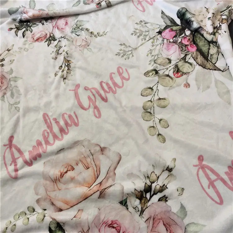Eine weiche cremefarbene Fleece-Baby-Wickeldecke, personalisiert, mit zarten rosa- und grünen floralen Illustrationen, wobei der Name Amelia Grace elegant in Roségold-Schrift gesetzt ist.