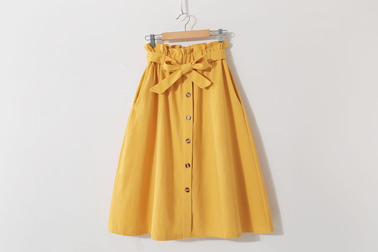 Une jupe midi en coton jaune vif présente une silhouette en ligne A à taille haute, une ceinture élastique froncée, un nœud en tissu assorti sur le devant et une rangée verticale de boutons métalliques au centre pour un port décontracté.