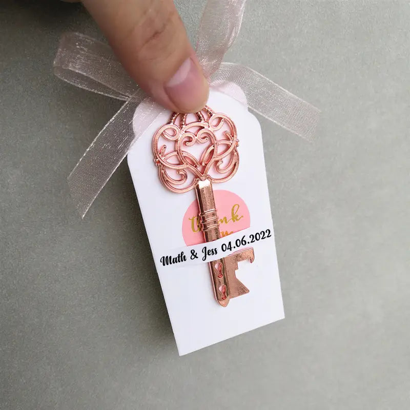 Abridor de botellas metálico con forma de llave ornamentada en oro rosa, con un lazo decorativo y una etiqueta blanca adherida, diseñado como recuerdo abridor de botellas para bodas personalizado.