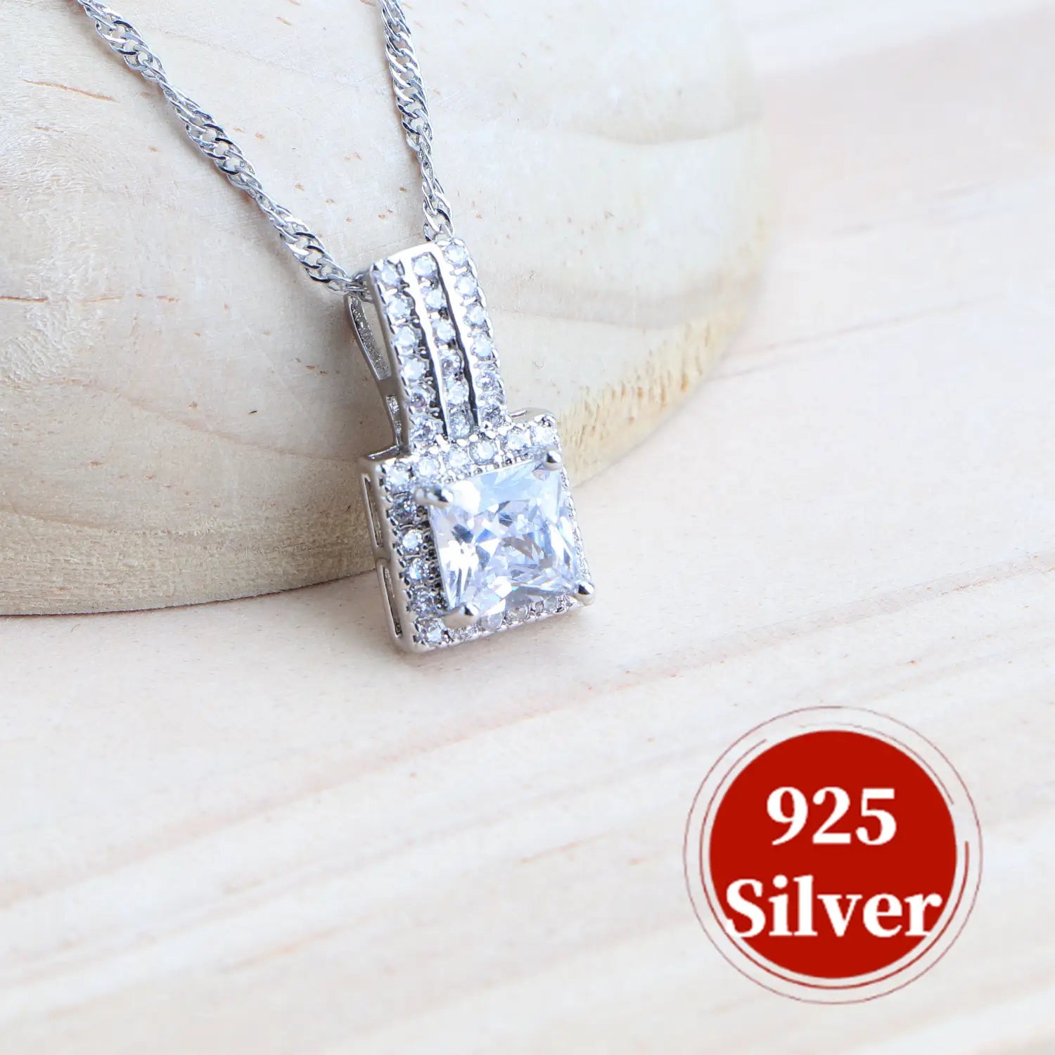 Un colgante de cristal de talla cuadrada, brillante, montado en un marco de plata de ley 925 con piedras más pequeñas a lo largo de los bordes, sujeto a una cadena de alambre trenzado, como parte de un juego de joyería nupcial de plata de ley 925 con cristales.