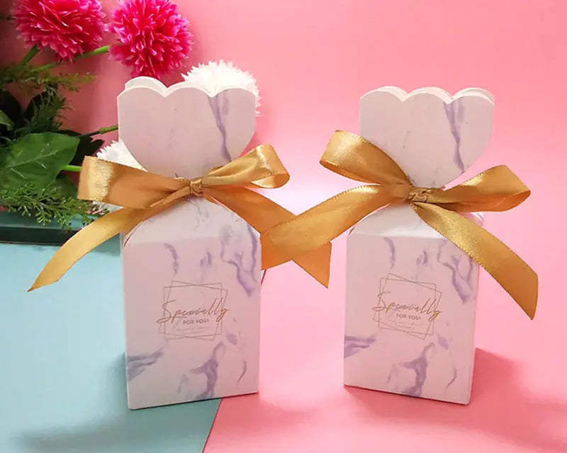 Blumige Hochzeits-Gastgeschenkboxen aus weißem, marmoriertem Material mit goldfarbenen Satin-Schleifen und mit „Speziell für Sie“ in eleganter Handschrift bedruckt.