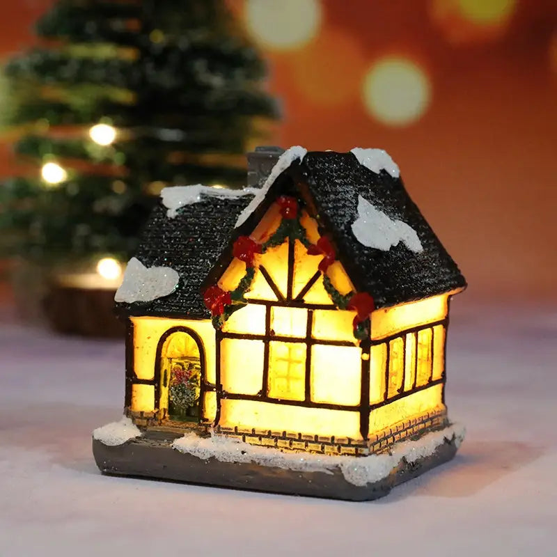 Une maison miniature du village de Noël à LED, illuminée, avec un toit en ardoises noires recouvert de neige blanche, des fenêtres jaunes lumineuses encadrées par des poutres en bois sombre, et décorée de baies rouges sur une guirlande verte, veilleuse à batterie.