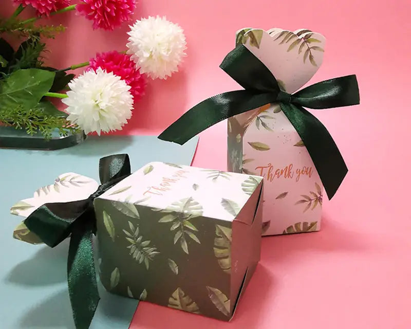 Zwei dekorative Blumengastgeschenkboxen, eine quadratisch und eine rechteckig, mit gedämpften Grünblattmustern auf weißen und rosafarbenen Hintergründen, gebunden mit glänzenden dunkelgrünen Satinbändern.