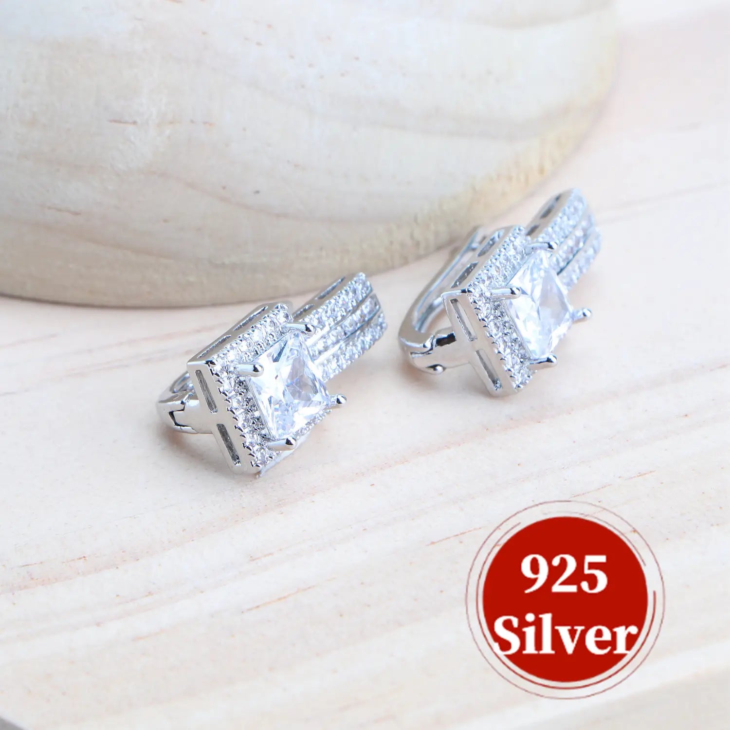 Un par de elegantes aretes tipo huggie de pedida nupcial de plata de ley 925 con cristales, que presentan una piedra central de talla princesa transparente rodeada de piedras pequeñas engastadas tipo pavé.