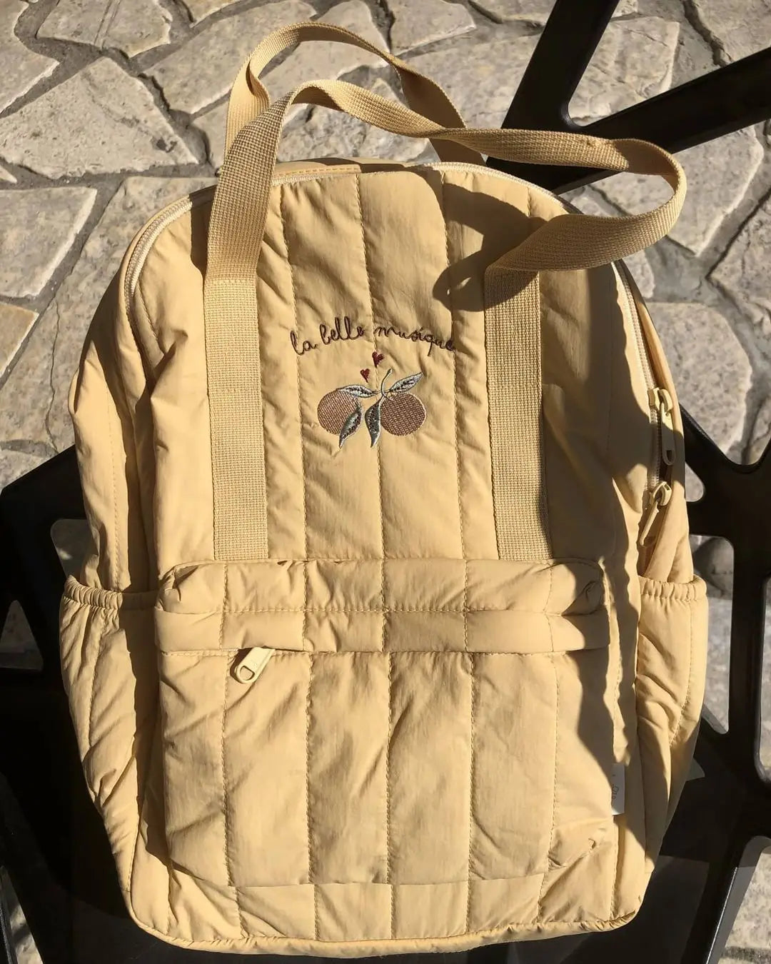 Ein weicher beiger gesteppten Kinder-Rucksack aus wasserfestem Material mit besticktem Obst und dem Schriftzug „la belle musique“ auf der Vorderseite, vorgesehen für den Schulgebrauch.