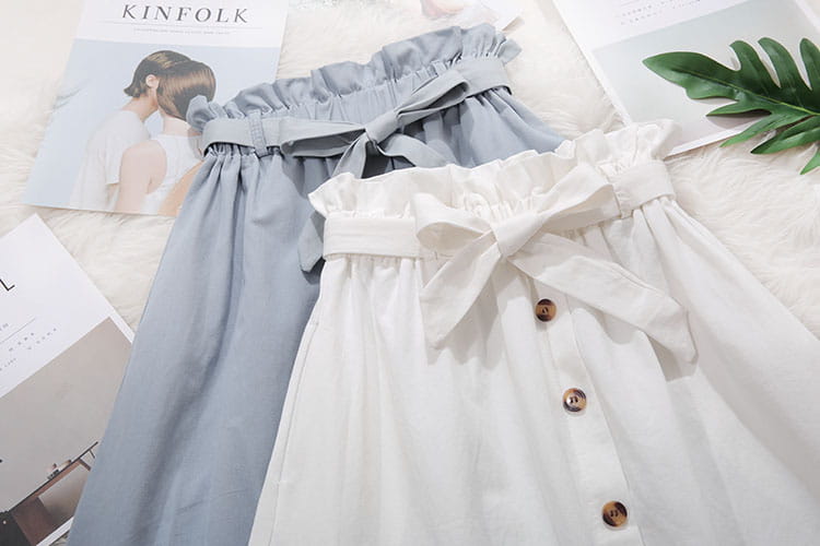 Une paire de jupes midi en coton bleu clair et blanc, à taille haute avec des bandes élastiques nouées en forme de nœuds, des boutons en bois sur le devant et des ourlets à volants, adaptées à un usage décontracté.