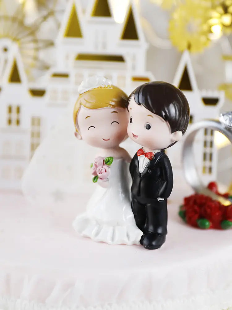 Cute cartoon-style wedding cake topper featuring a gold castle acrylic decor with a blonde bride in white and a groom in black tuxedo with red bow tie.