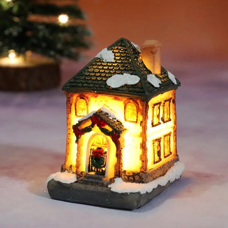 Une maison miniature du village de Noël à LED, éclairée chaleureusement, avec un toit en tuiles vertes et des détails blanchis par la neige, ainsi que des guirlandes festives autour de l’entrée, veilleuse à batterie.
