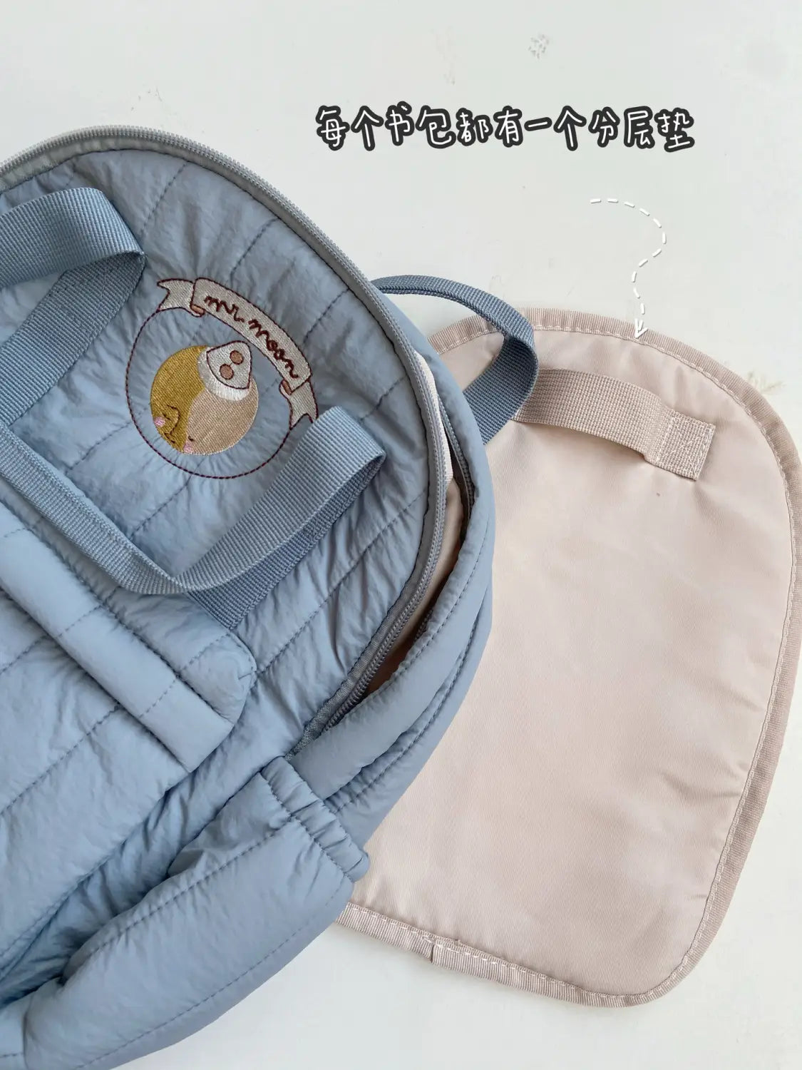 Hellblauer gesteppt wasserfester Kinder-Rucksack mit einer runden bestickten Applikation mit einem Cartoon-Charakter und einem Banner, geeignet für die Schule.
